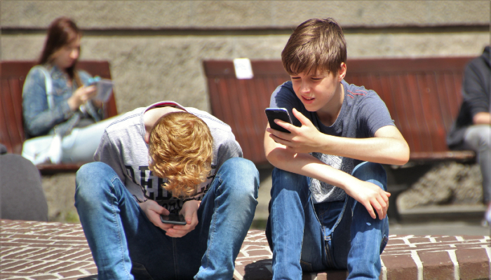 Colour photo of 2 boys sitting and looking at their phones. A woman in the background is also on her phone.