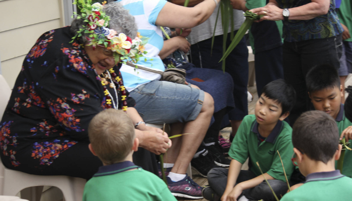 Colour photo of a group of school students listening to a woman doing a demonstration. It was part of a pacifica weaving workshop.