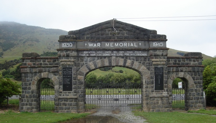 Colour photo of a WW2 memorial at Little River, Aotearoa NZ, made of stone with arches and wrought iron gates. The inscription includes the years '1939' and '1945' and says 'Greater love hath no man than this, that a man lay down his life for his friends'.