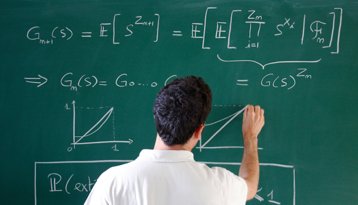 Colour photo of a man writing different equations in chalk on a green blackboard.