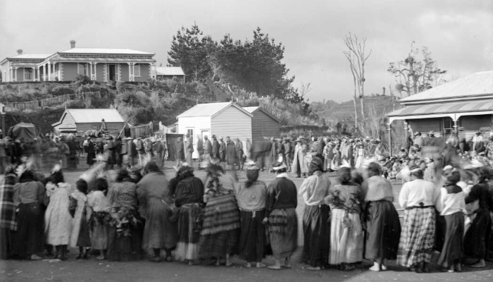 Black and white photo of a group and buildings at Parihaka Pā. In the foreground, people are standing side by side.