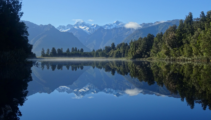 Colour photo of Te Ara Kairaumati (Lake Matheson) in Aotearoa NZ. The lake reflects the surrounding trees, Aoraki (Mount Cook) and other peaks.