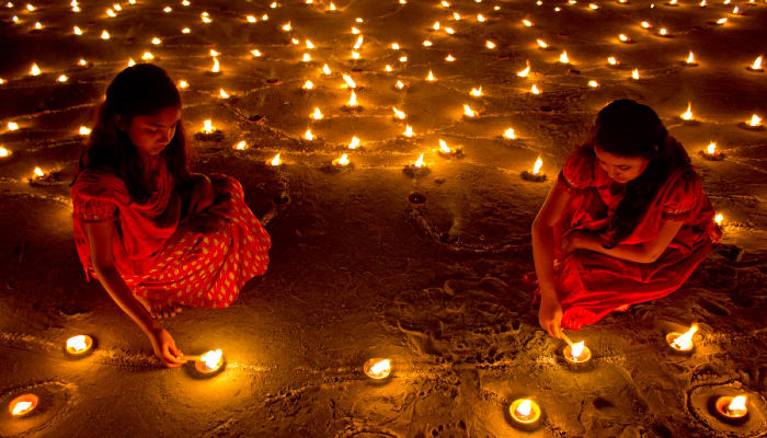 Colour photo of 2 girls lighting candles and diyas (clay lamps). They are surrounded by diyas that have already been lit and are wearing saris.