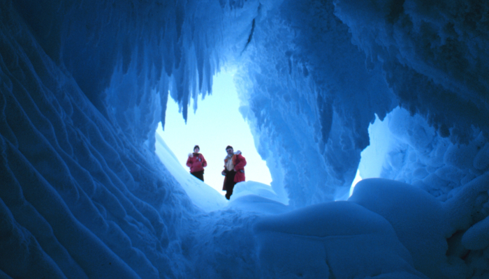 Colour photo from the Erebus Ice Tongue in Antarctica. It shows 2 people looking down a hole in an ice cave.