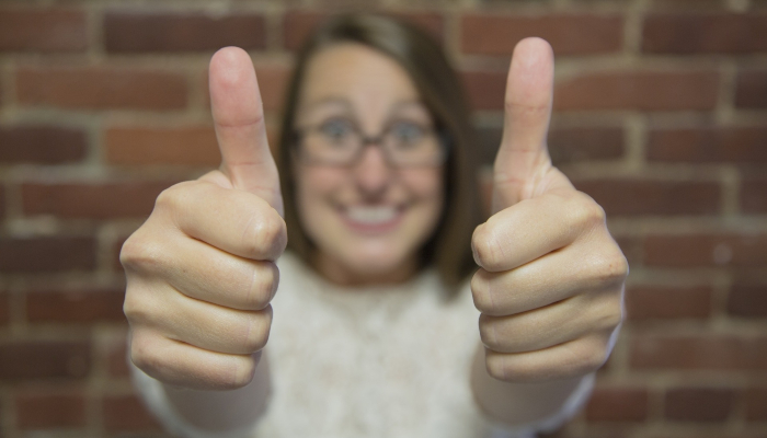 Colour photo of a smiling woman holding both arms outstretched, with fists closed and thumbs pointing upwards to convey approval. 