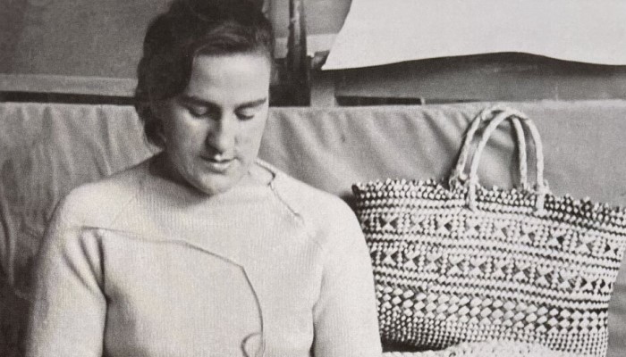 Black and white photo of Cath Brown sitting on a sofa weaving. There is a kete (woven basket) beside her.