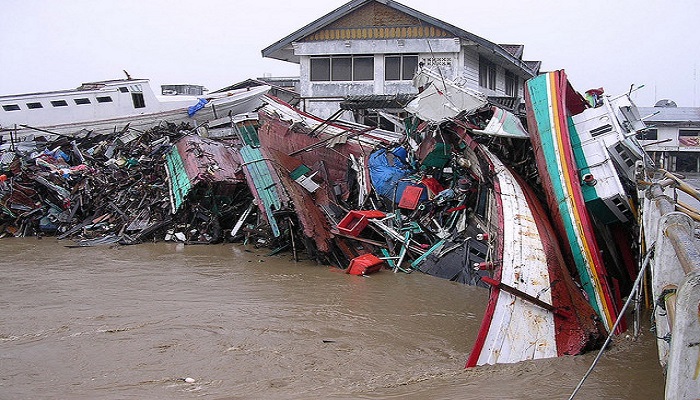 Colour photo showing smashed boats piled up next to a building, with brown water all around.