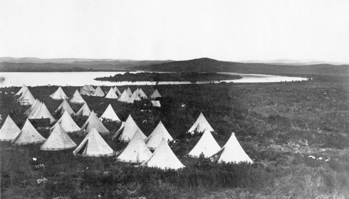 1863 black and white photo of a group of bell tents near the Waikato River. The tents are in the foreground to the left of the image, in the distance the river and a small island are visible.