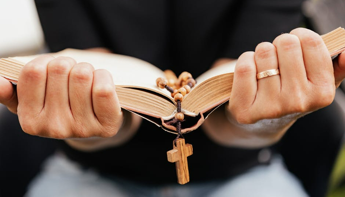 Colour photo of a person holding a book open with a rosary placed inside and a cross hanging over the edge.