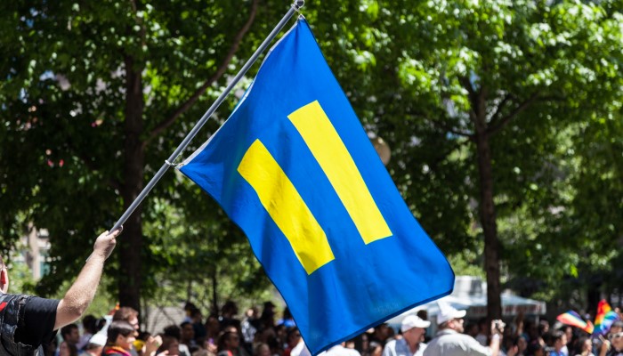 Colour photo of a flag being waved at a parade. The flag has the Human Rights Campaign logo, a blue background with yellow equals sign.