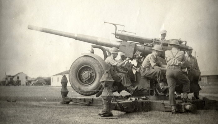 Black and white photo from WW2 showing soldiers operating an anti-aircraft gun in the middle of a field.