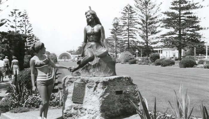 1967 black and white photo of the statue 'Pania of the Reef' on the Marine Parade, Napier. A woman stands beside the statue.