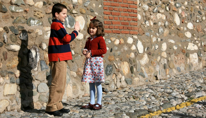 Colour photo of a boy and a girl standing in front of a brick wall facing each other. The boy is pointing to his other hand.