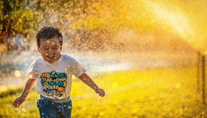 Colour photo of a child smiling and playing while being sprayed with water.