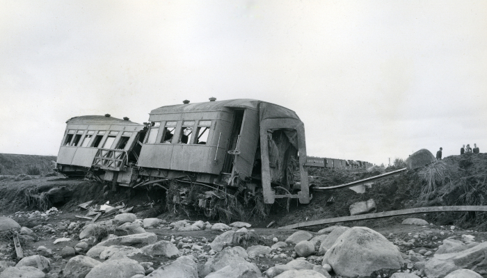1953 black and white photograph of Tangiwai showing a wrecked train carriage sitting on the edge of a stony riverbed.