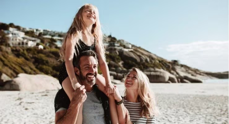 A couple walk on the beach, the man carrying their girl child offspring on his shoulders