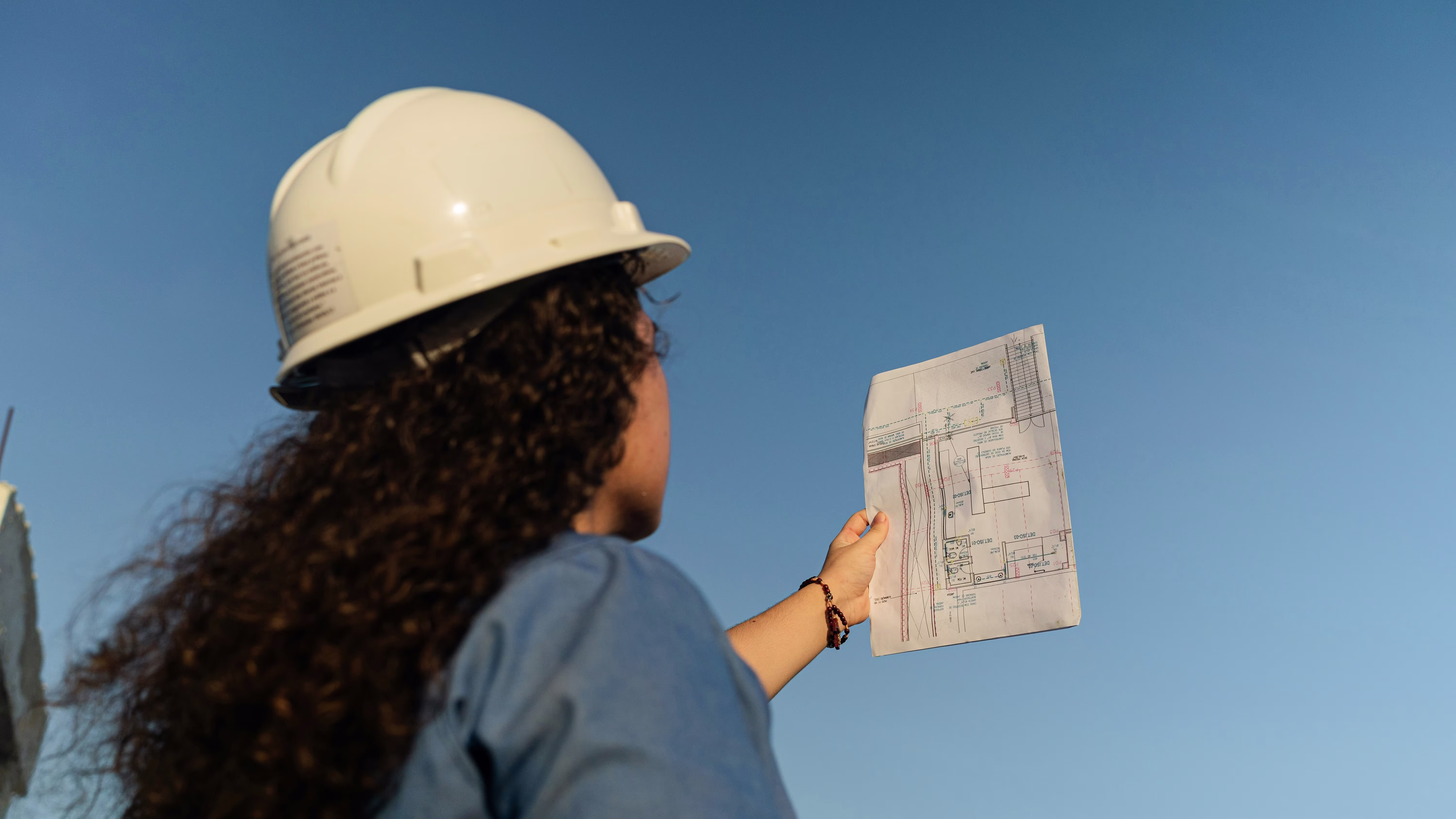 A woman wearing a white hard hat reviews construction blueprints outdoors against a clear blue sky.