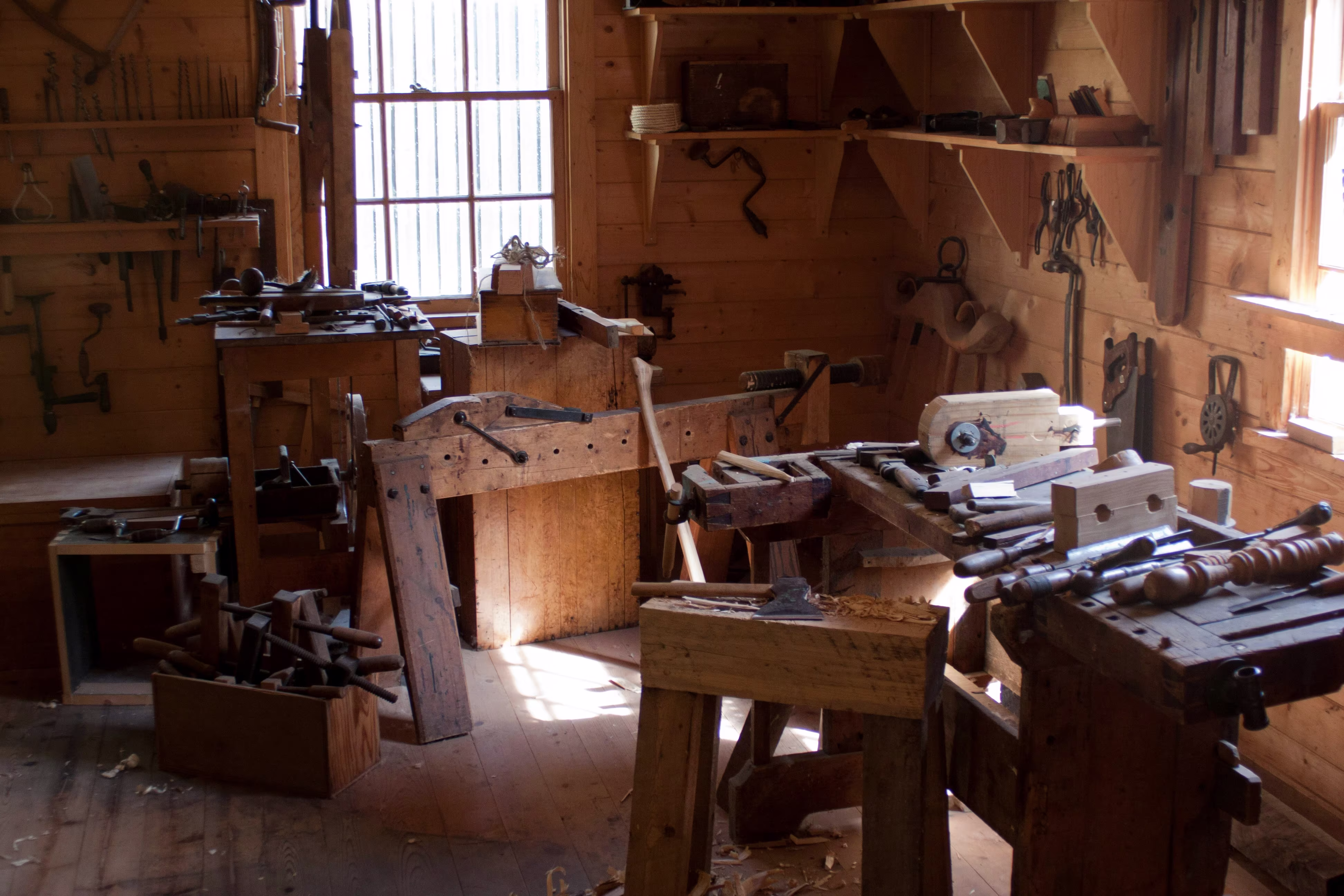 A rustic wooden carpentry workshop with workbenches, hand tools, chisels, and saws mounted on warm brown timber walls.