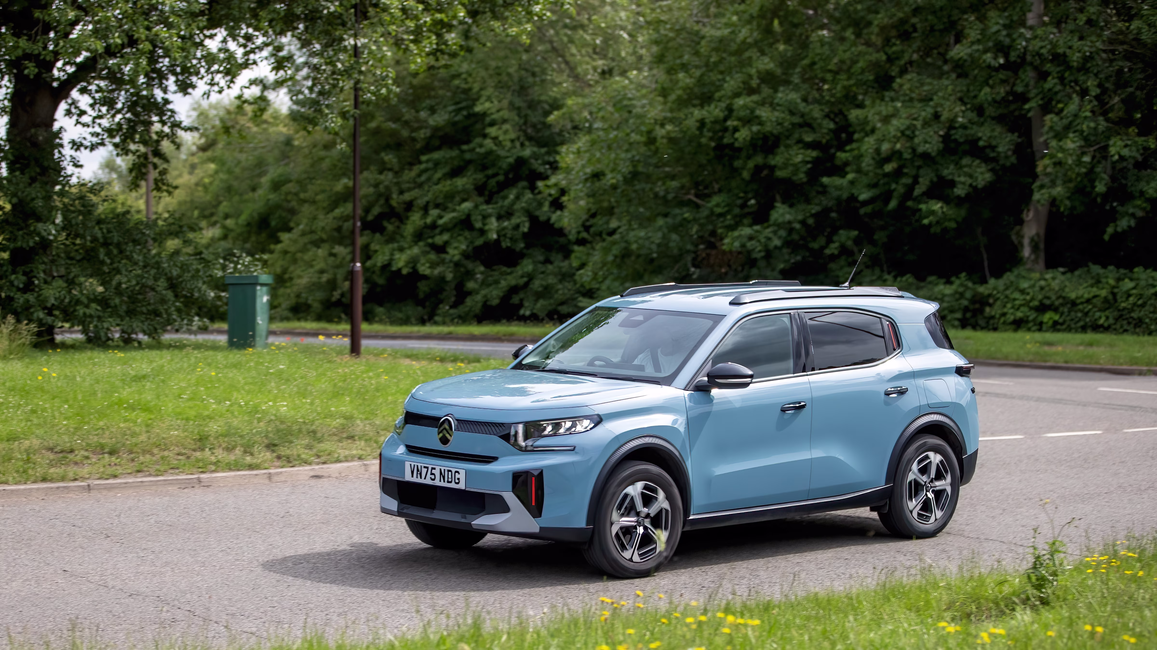 A light blue Citroën ë-C3 SUV on a road, surrounded by lush green trees and grass on a cloudy day.