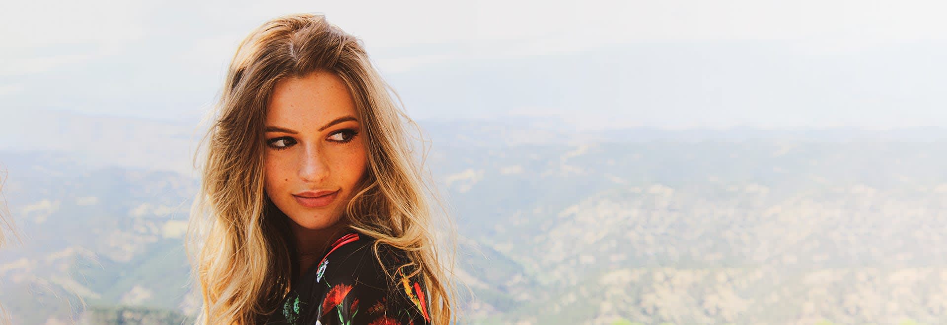 A photo of a woman with long blonde hair looking on the side - mountains in the background