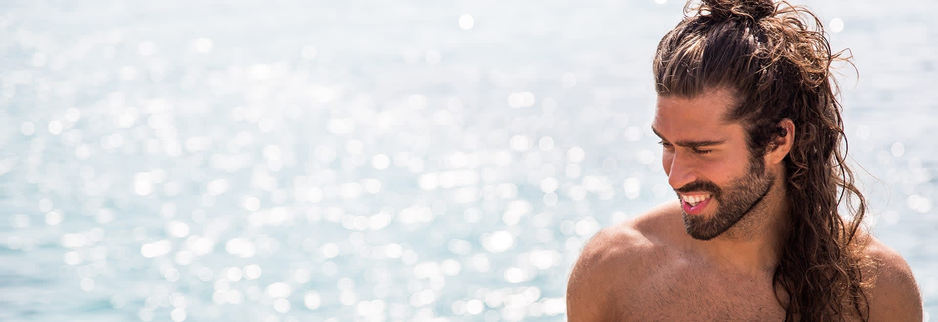 A photo of a man with long dark hair pinned up standing stand in front of the ocean