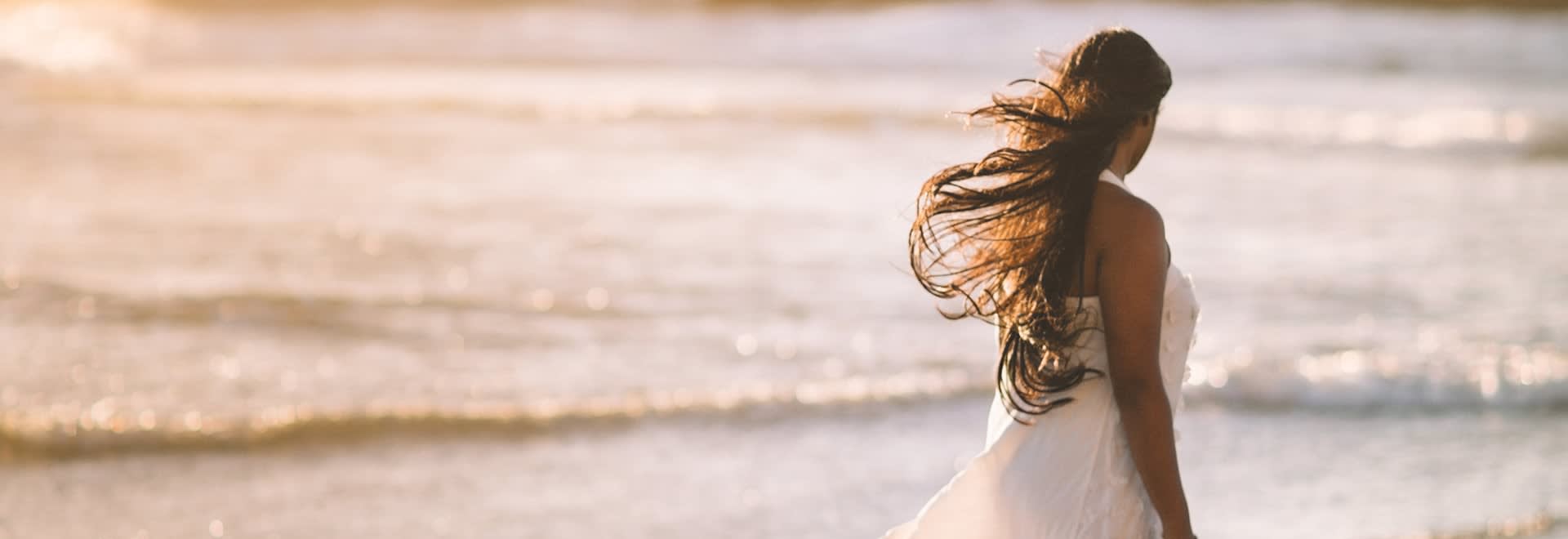 A woman with her long dark hair blown up by the wind standing backwards to the camera on the beach