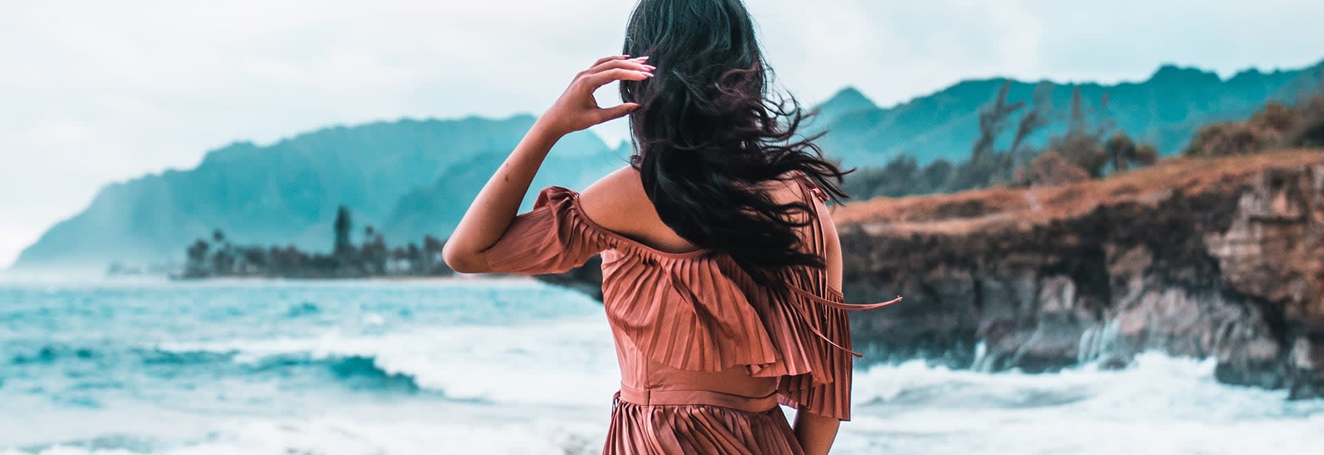 A photo of a woman with long dark hair blown up by the wind looking ahead standing backwards to the camera - the ocean in the background