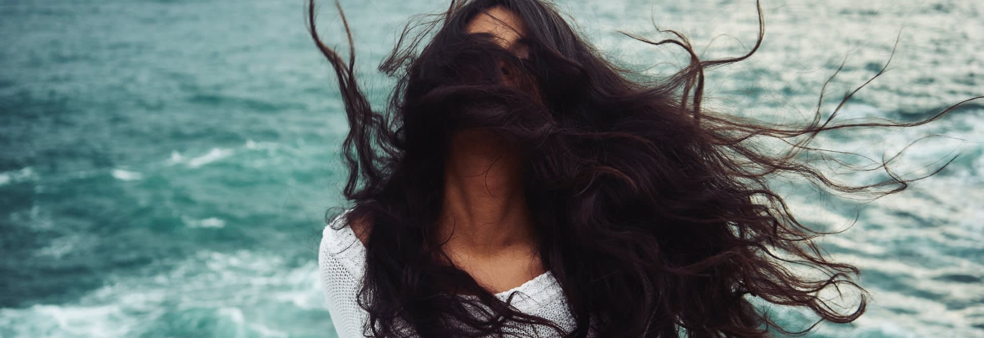 A photo of a woman on the coast with long dark hair blown by the wind