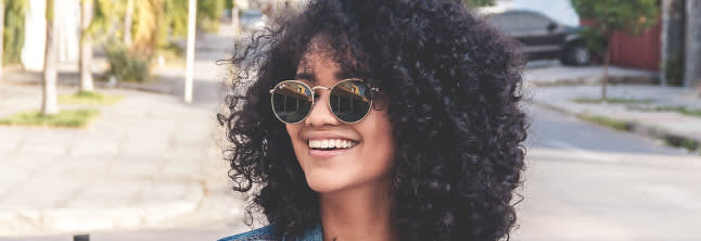 A young woman with mid-lenght curly hair wearing sunglasses and smilling, the street in the background