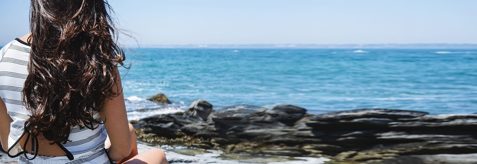 Picture of a woman with brown hair sitting on the shore and looking at the ocean