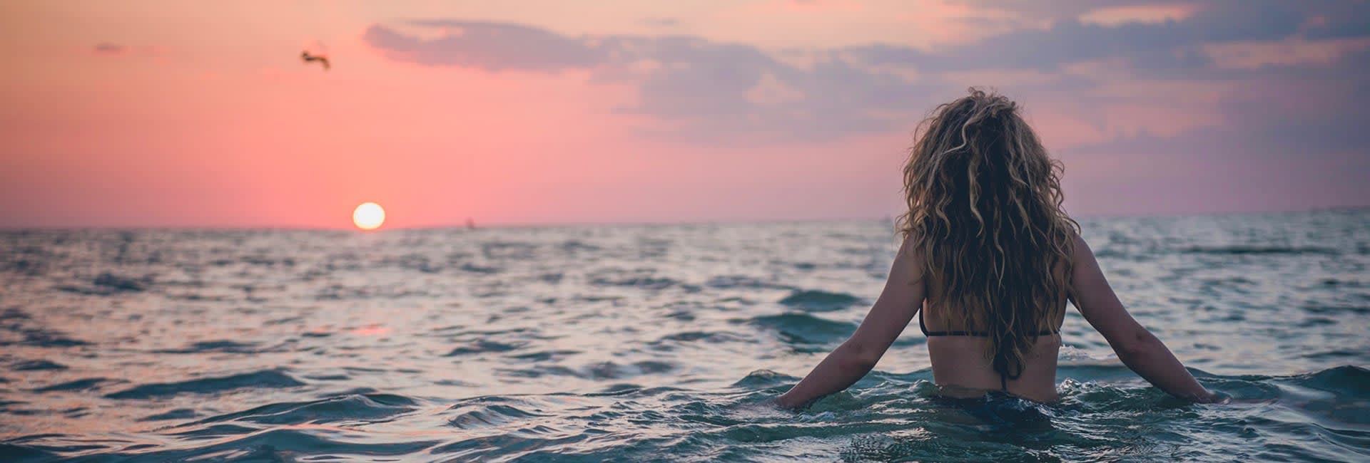 A photo of a woman with long dark-blonde curly hair in the ocean going in the direction to a sunset