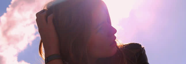 A photo of woman touching her head with a sunlight hitting the camera and a sunset in the background