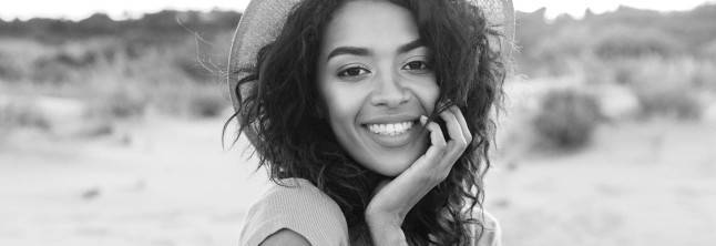 A black and white photo of a woman with long curly dark hair standing on a beach, smiling and touching her face