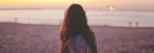 A photo of a woman with long dark hair looking ahead at the ocean standing on a beach backwards to the camera