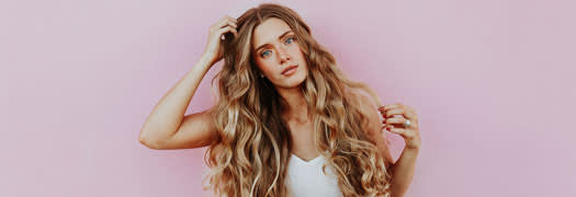 A photo of a woman with long fair wavy hair looking at the camera, pink wall in the background