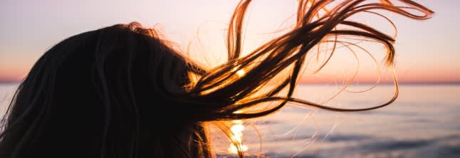 A woman with her hair blown up by the wind standing backwards to the camera with the sunset in the background