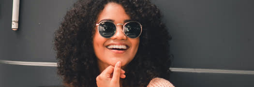 A young woman with mid-lenght curly hair wearing sunglasses and smilling, the wall in the background