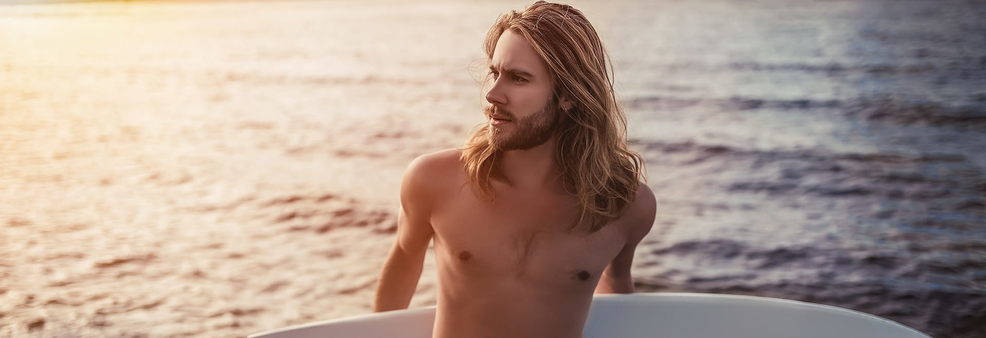 A photo of a man with long blonde hair holding a surfboard on the beach 
