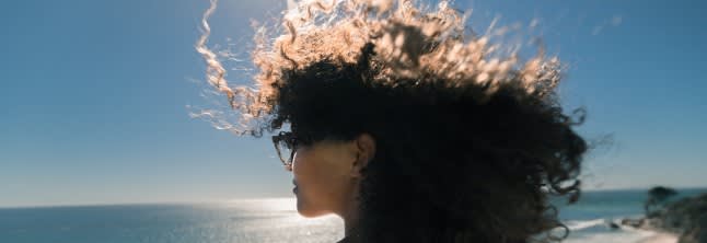 A photo of a woman with curly dark hair blown up by the wind looking ahead and standing backwards to the camera on the clear-blue sky background