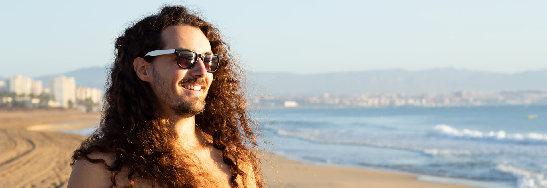 A young man with long curly hair standing on a beach with sunglasses on