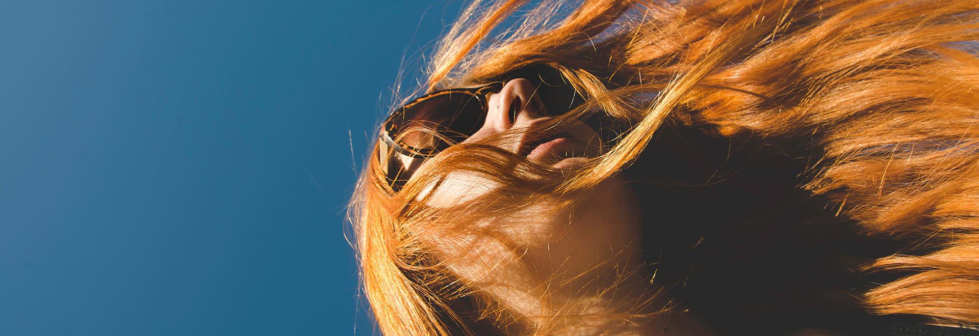 A photo of a woman with long red hair wearing sunglasses on the clear blue sky background - a shot from below 