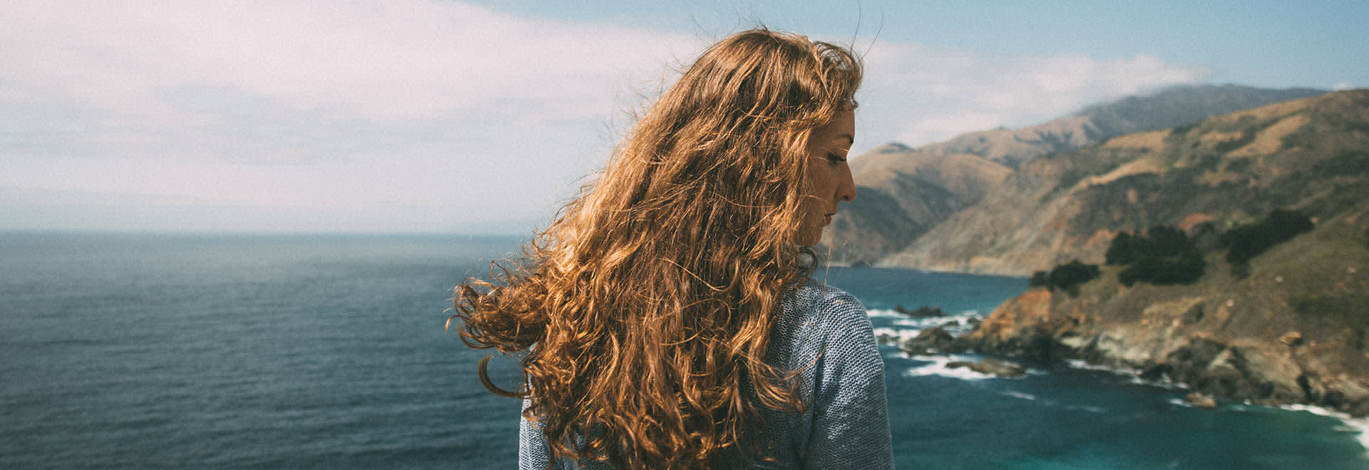 A photo of a woman with long wavy red hair looking over her shoulder on the side - mountains and ocean in the background