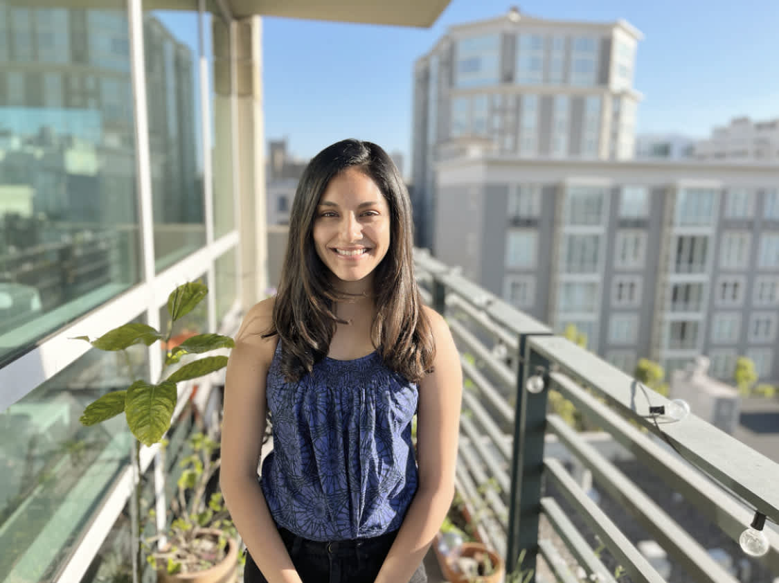 Aastha, a young woman with shoulder-length brown hair and light brown skin, wearing a sleeveless purple top. She stands on a balcony in San Francisco with a city view behind her. 