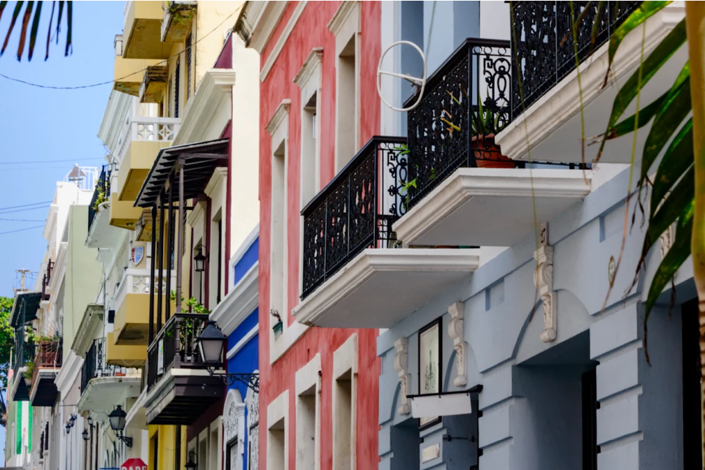 Colorful buildings with ornate black balconies and plants, viewed from the street on a sunny day.