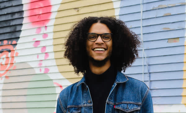 Jacob, a young man with curly brown hair and medium-brown skin, wearing black-rimmed glasses and a denim jacket over a black shirt. Behind him is a mural of colorful circles and shapes.