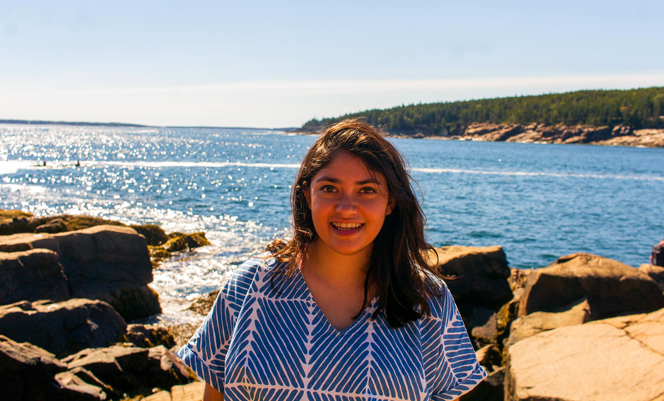 Runi, a young woman with light brown skin and shoulder-length brown hair, wearing a short-sleeved top with diagonal blue stripes. Behind her are water, rocks, and shoreline.