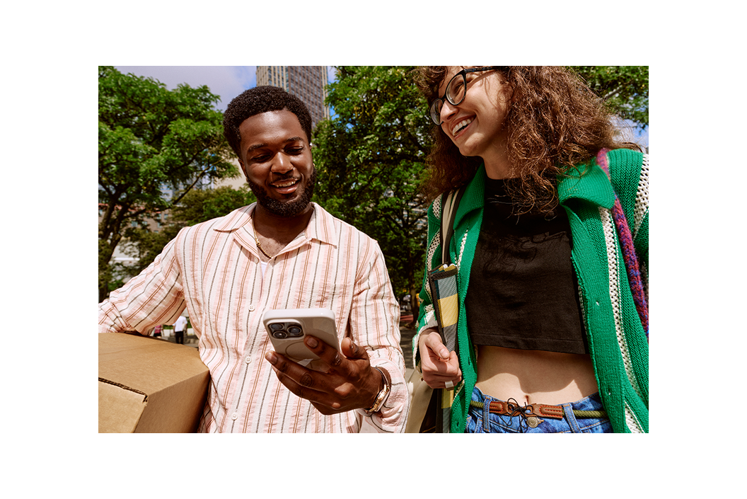 Two people outdoors smiling, one holding a phone and a box, the other wearing glasses and a green sweater.