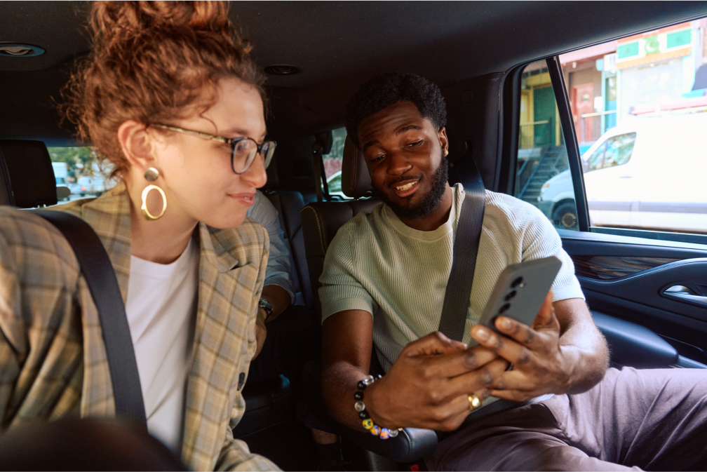 Two passengers in a car, both wearing seatbelts, looking at a smartphone and smiling.