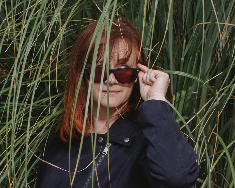 Kat, a woman with light skin and shoulder-length red hair, wears a stylish black jacket and peers sideways over her sunglasses. Behind her is a background of giant grasses.