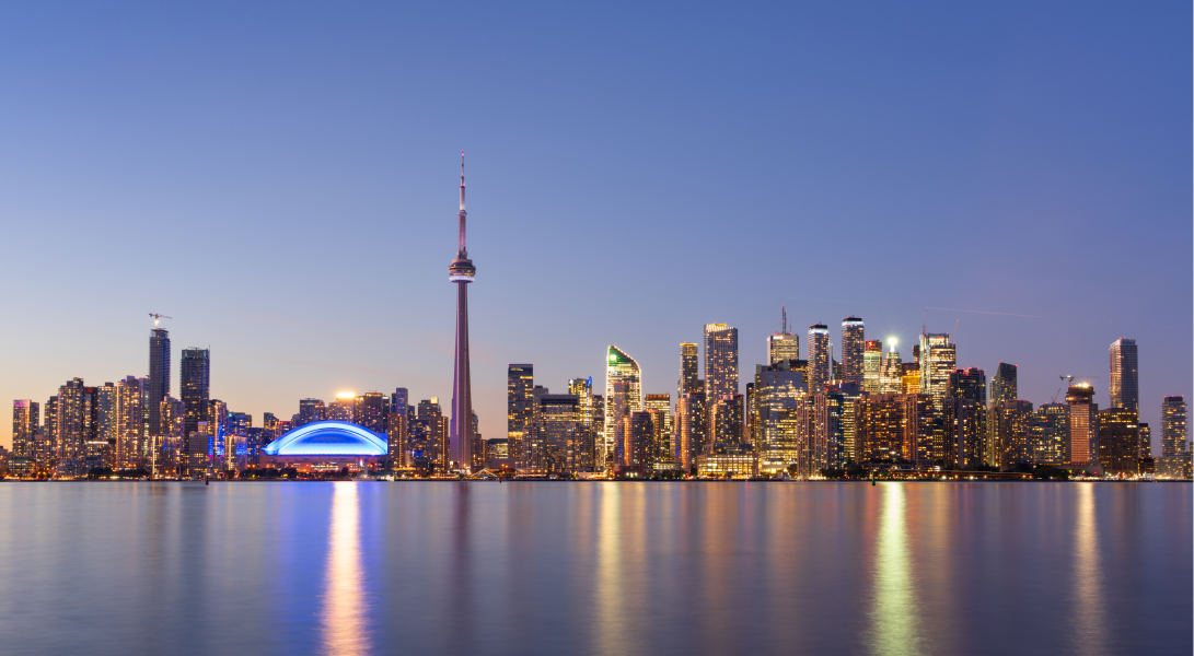Toronto skyline at dusk with city lights, CN Tower, and blue-lit stadium reflected on calm water.